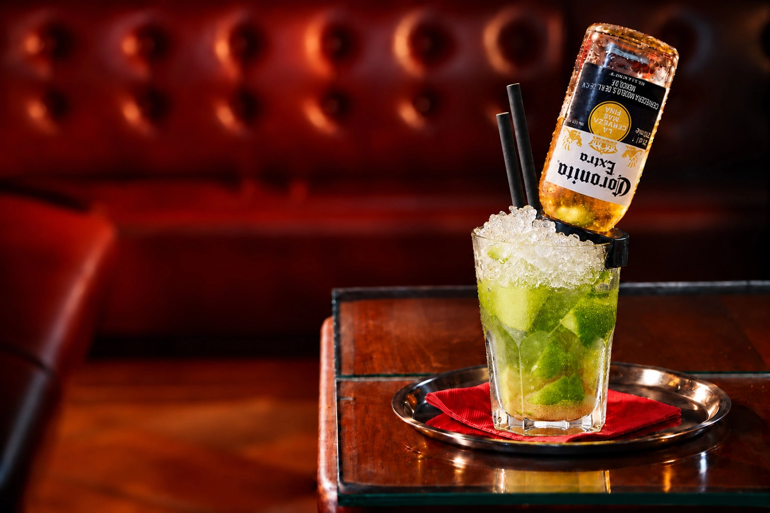Coronita Pirinha mit Limetten, Crushed Ice, umgedrehter Coronita-Flasche und schwarzen Papierstrohhalmen